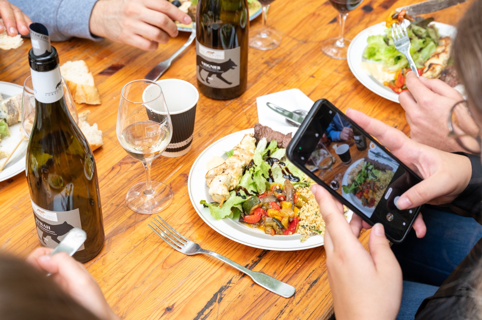 Bouteilles de vin et assiettes du brunch posées sur une table. Une personne est en train de prendre une photo avec un téléphone.
