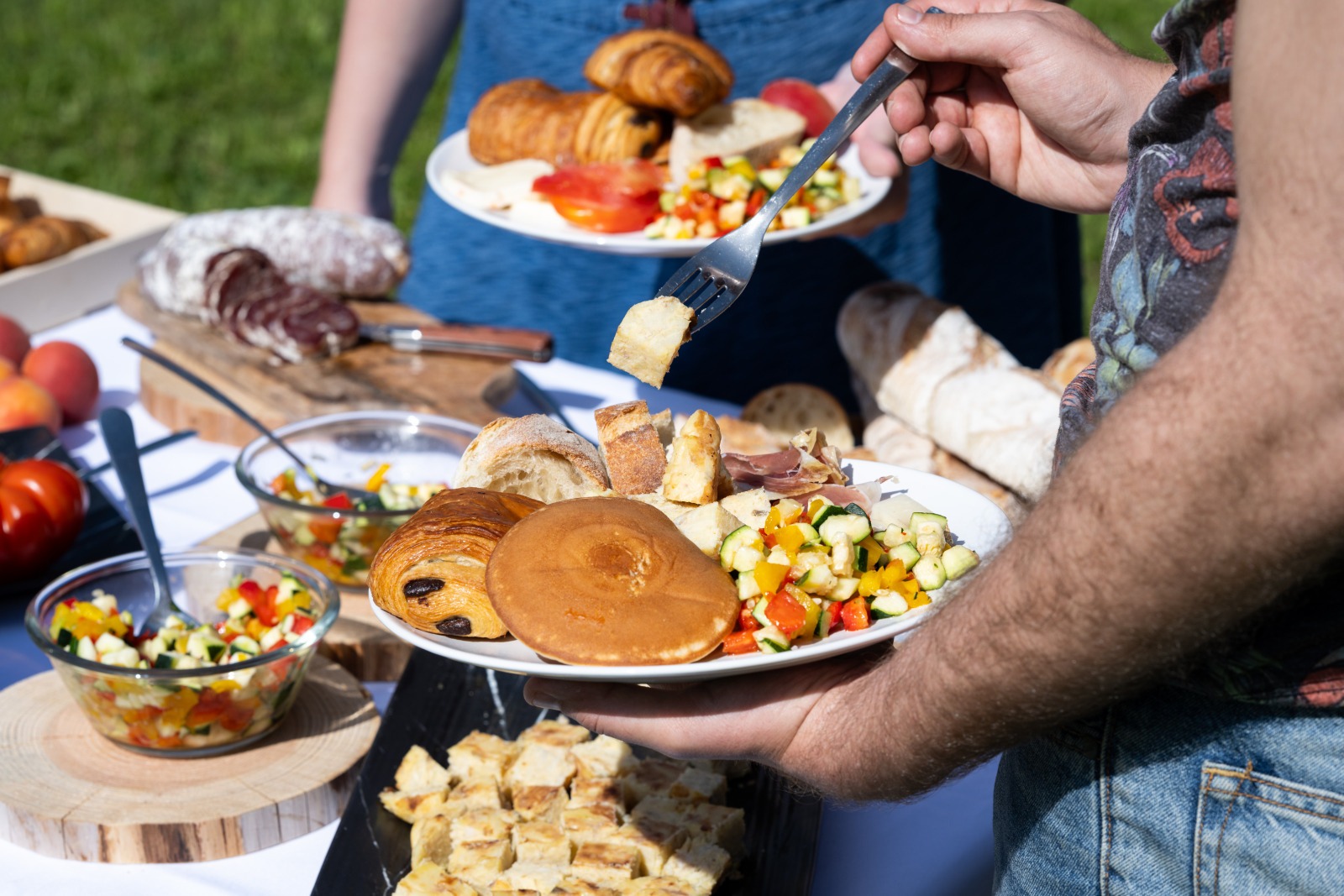 Brunch Vigneron installé sur une table avec une nappe blanche posée au milieu de l'herbe. Une personne est en train d'attraper de la nourriture avec une fourchette.